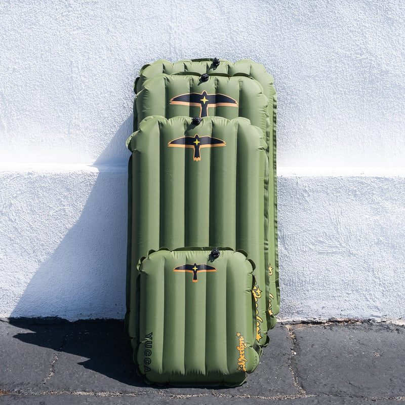 Four olive green Yucca Merlyn inflatable surf mats in different sizes stacked against a white textured wall outdoors. Each mat features the black and gold bird logo and vertical air chambers. Sizes shown compare Mini, Junior, Standard, and XL options for size and volume differences.
