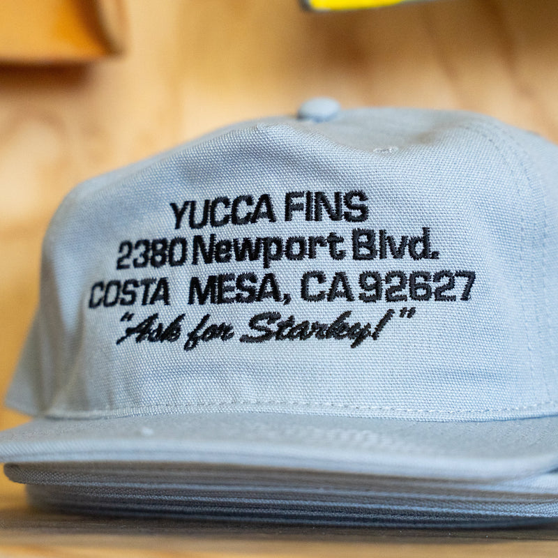 A light blue flat-brim baseball cap with black embroidered text reading 'YUCCA FINS 2380 Newport Blvd. COSTA MESA, CA 92827 