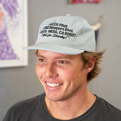 A smiling man wearing a light gray baseball cap with black embroidered text reading 'HUCCA FINS 2580 Newport Blvd. Costa Mesa, CA 92627 Ask for Stanley'.