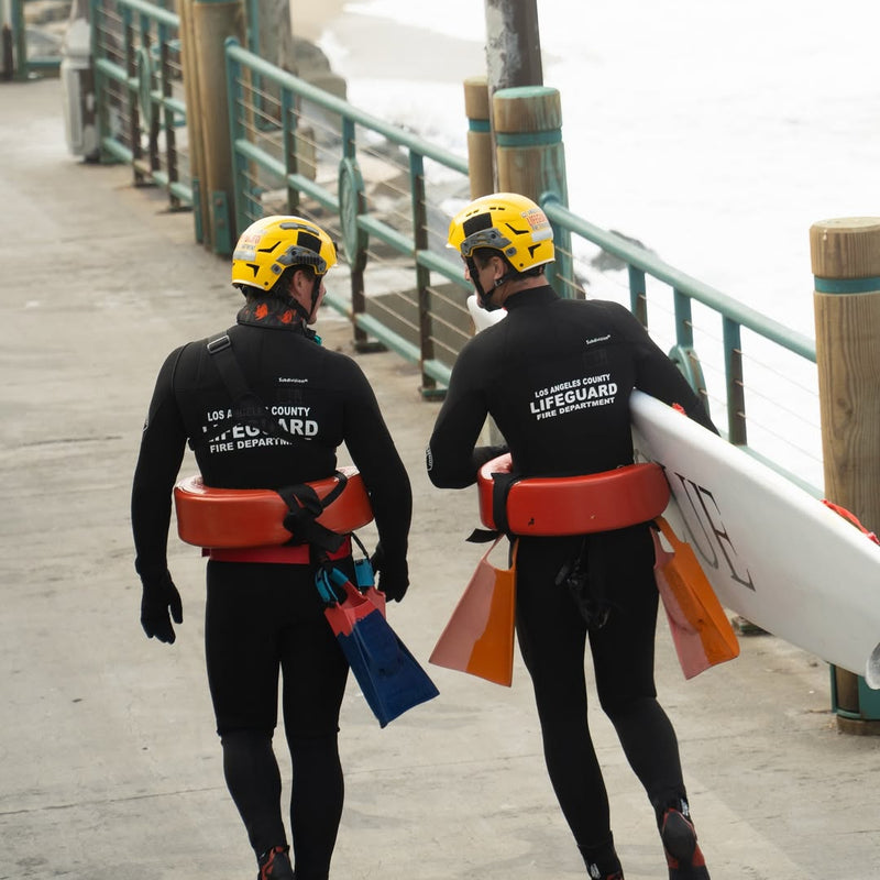 Two lifeguards in wetsuits and helmets walking on a wooden pier with surfboards and rescue equipment.